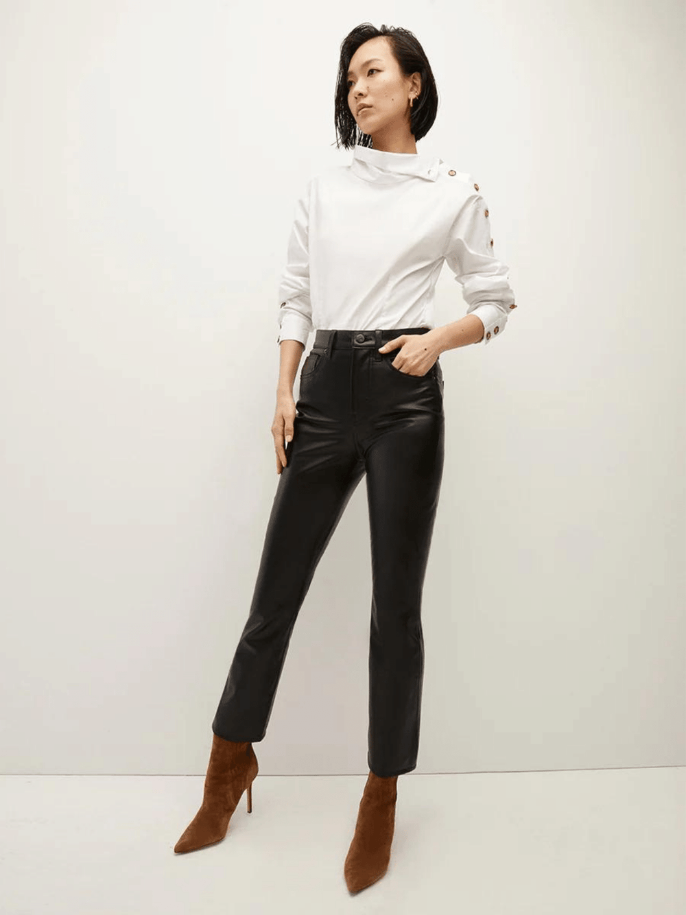 Ein Model trägt eine stilvolle weiße Bluse mit einem asymmetrischen Kragen und einer einzigartigen Knopfleiste entlang des linken Ärmels, kombiniert mit schwarzen Lederhosen.