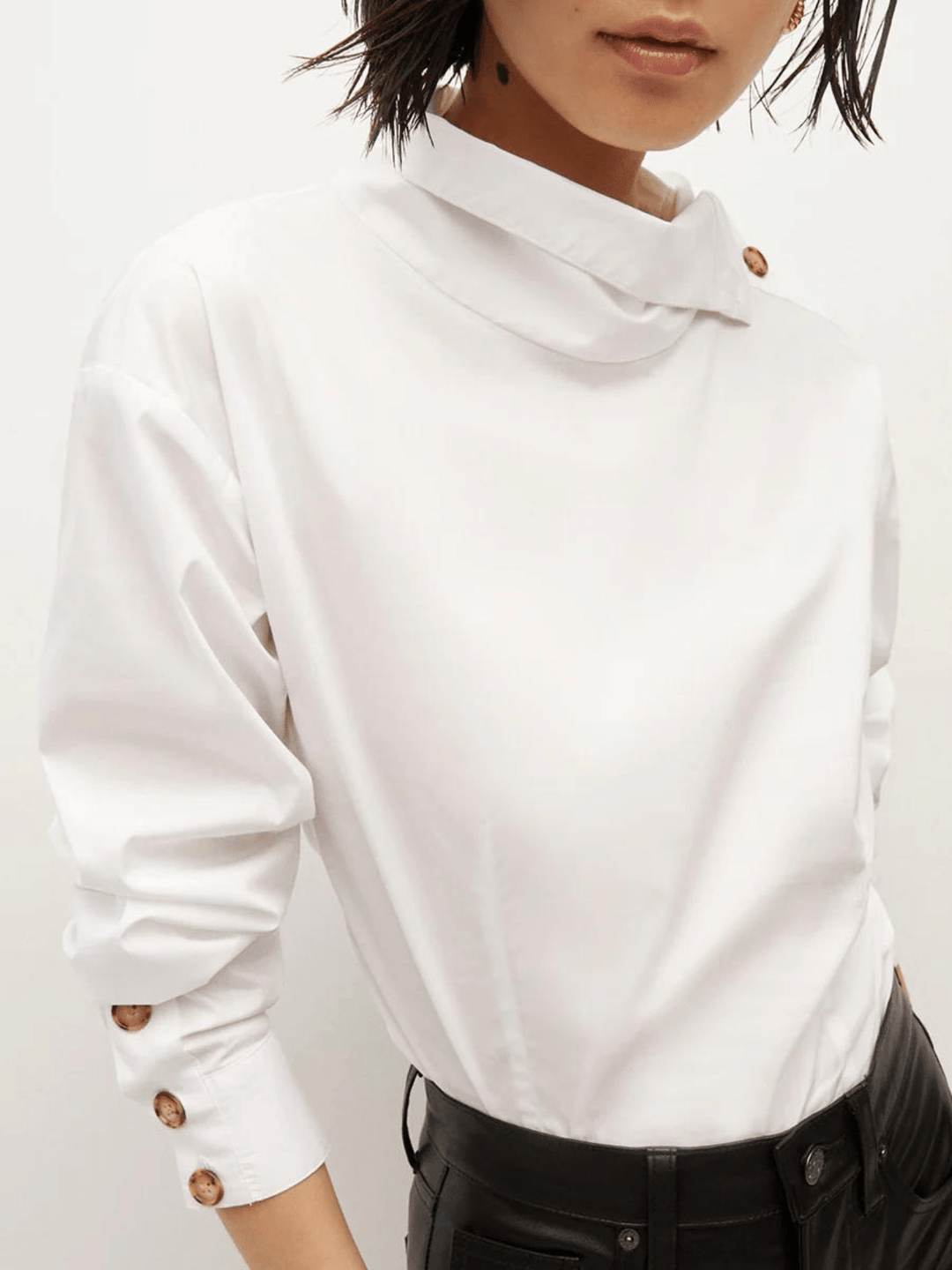Ein Model trägt eine stilvolle weiße Bluse mit einem asymmetrischen Kragen und einer einzigartigen Knopfleiste entlang des linken Ärmels, kombiniert mit schwarzen Lederhosen.